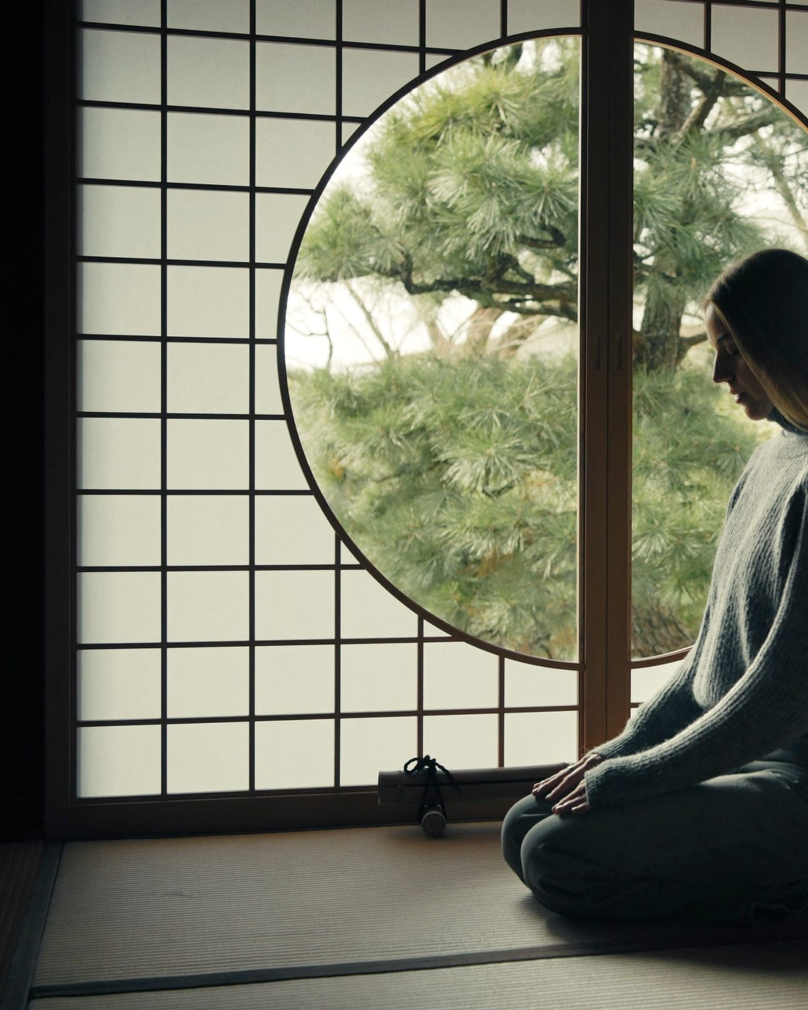 靜坐者側影，面向日式圓窗外的松——身體先安靜下來，意識才抵達。
